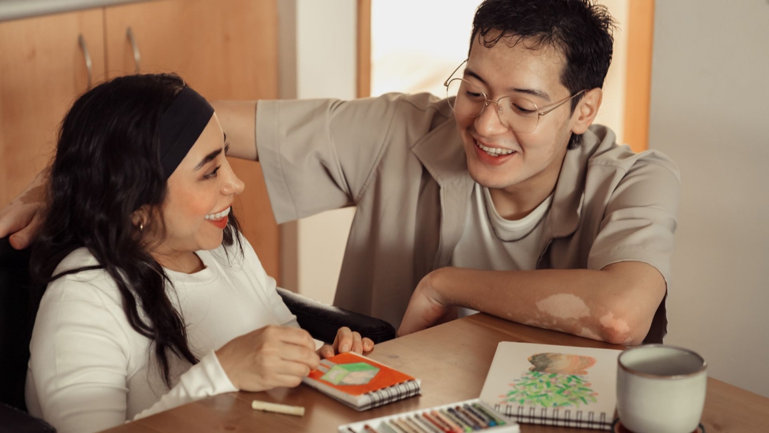 Two people sitting at a table together, artwork in front of them, smiling at each other.