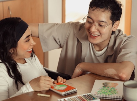 Two people sitting at a table together, artwork in front of them, smiling at each other.